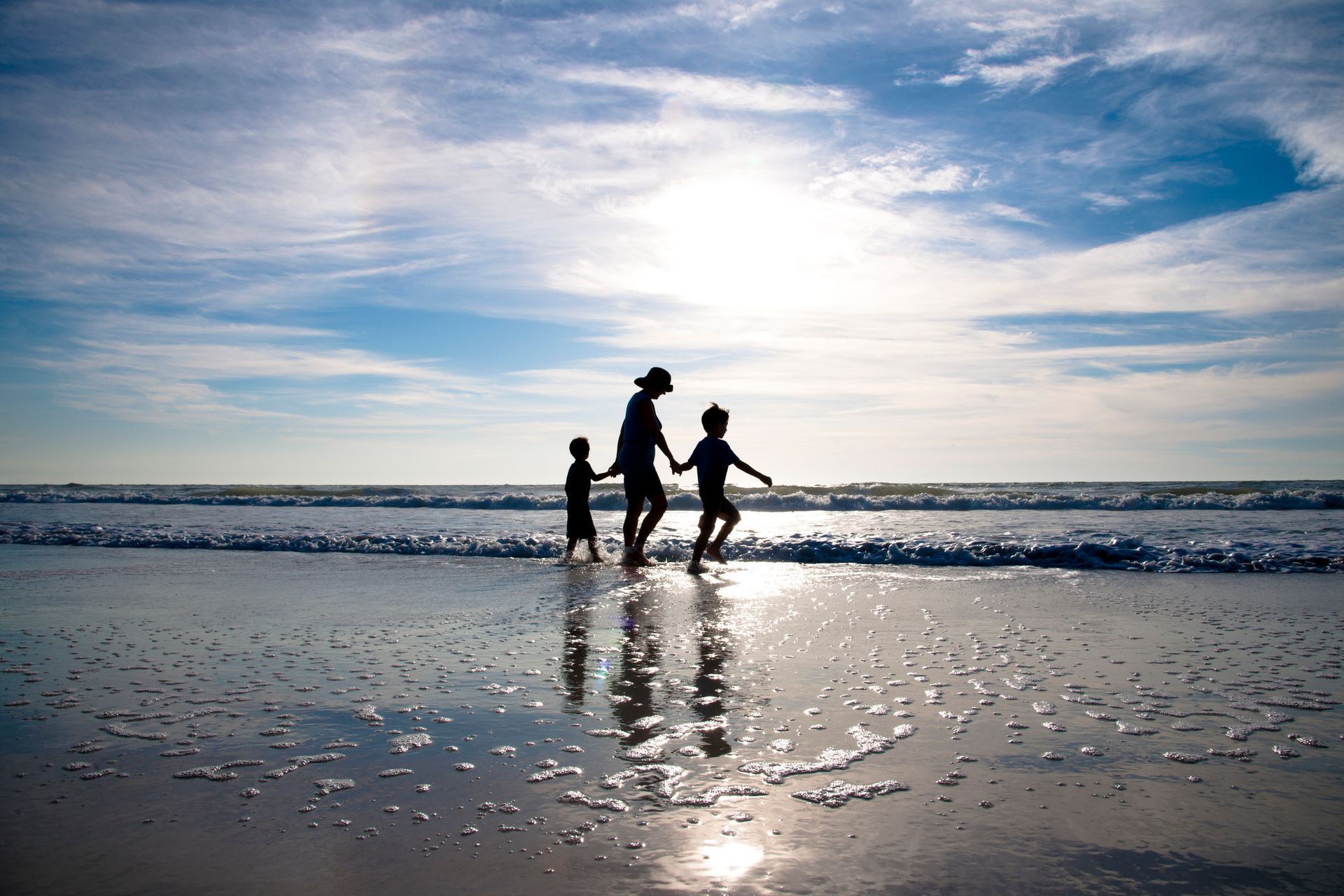 Three people holding hands and running on a sunny beach with reflections on wet sand