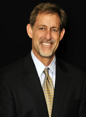 Meet Our Attorney, Drake Ozment Smiling man in a dark suit, white shirt, and patterned tie against a black background