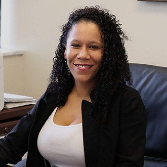 Jeanice Colon Smiling woman seated at a desk in an office, wearing a black blazer and white top.