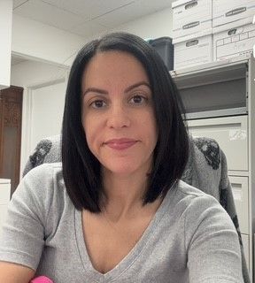 Jessica Abrue Woman seated at a desk in a gray top, smiling in an office with shelves and storage boxes behind her.