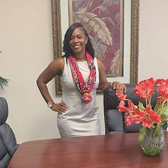 Deanna Simmons Woman in a gray dress stands smiling beside a conference table with red flowers in a vase.