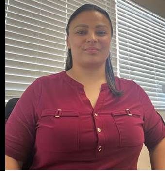 Evette Morales Woman in a burgundy top standing indoors in front of window blinds, looking at the camera.