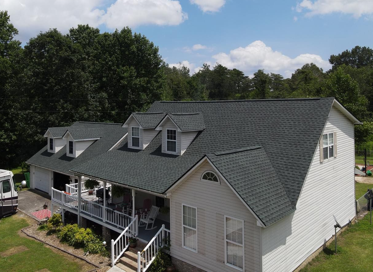 A white two-story house with a grey shingled roof, dormer windows, and a front porch, set in a sunny, green landscape.