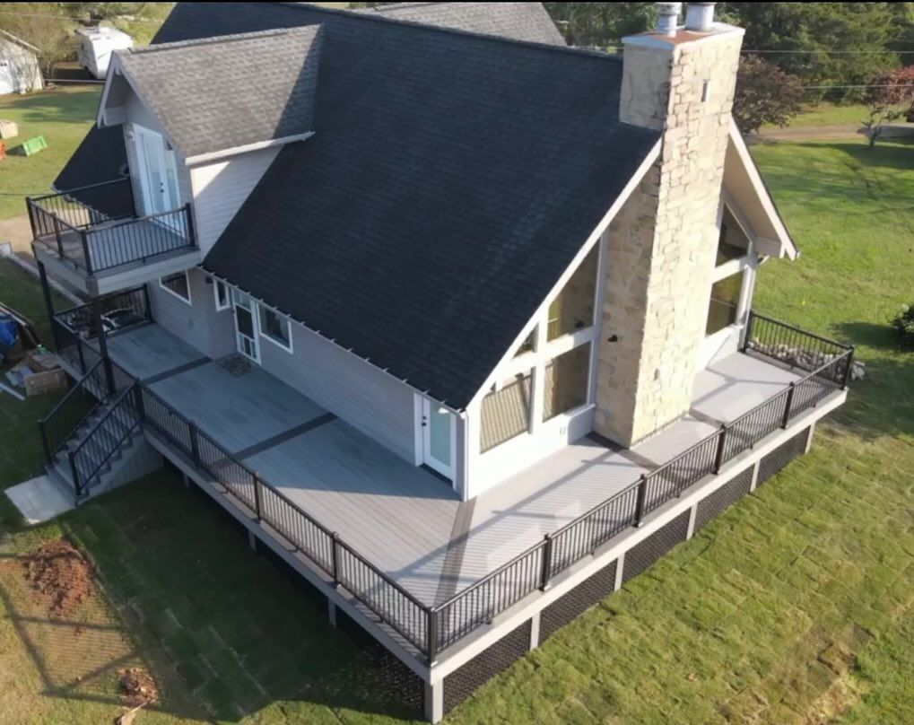 An aerial view of a gray house with a black roof, large deck, and a stone chimney, situated on a green lawn.