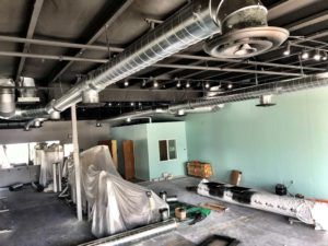 Interior of a commercial space under construction, showing ductwork, lighting, and covered furniture against a pale green wall.