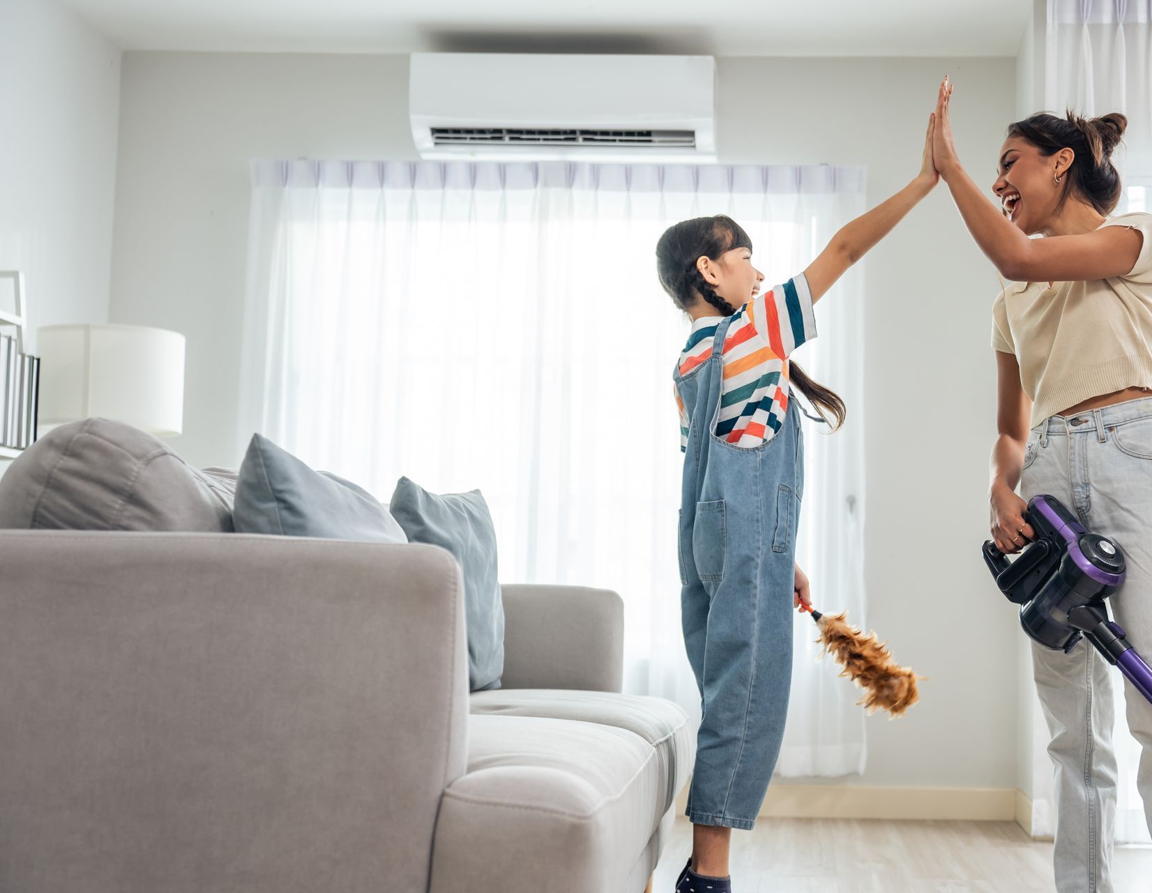 Woman and child high-five in a living room after cleaning. Child holds a duster, woman holds a vacuum.