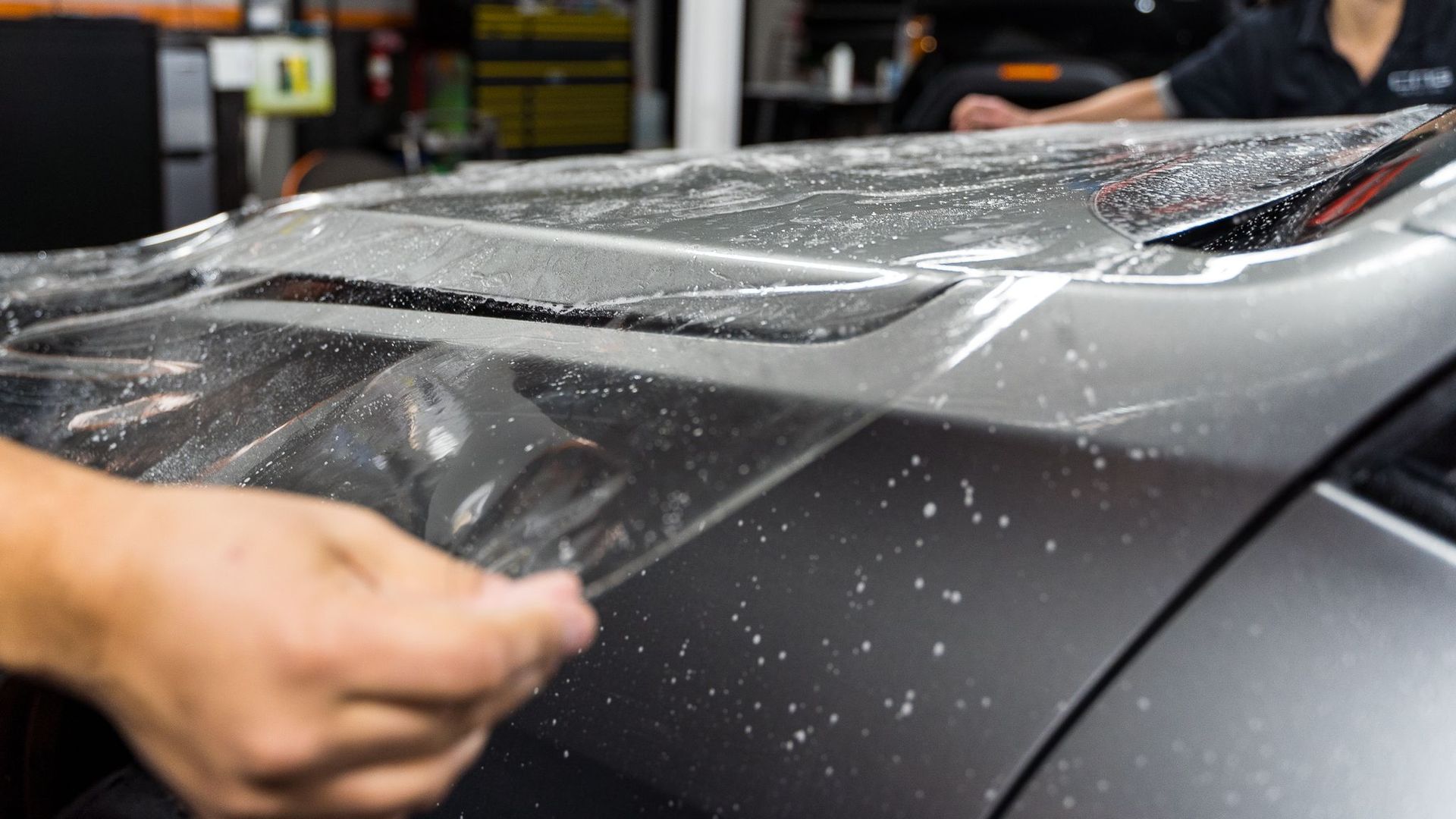Applying clear protective film to the roof of a gray car in a shop.