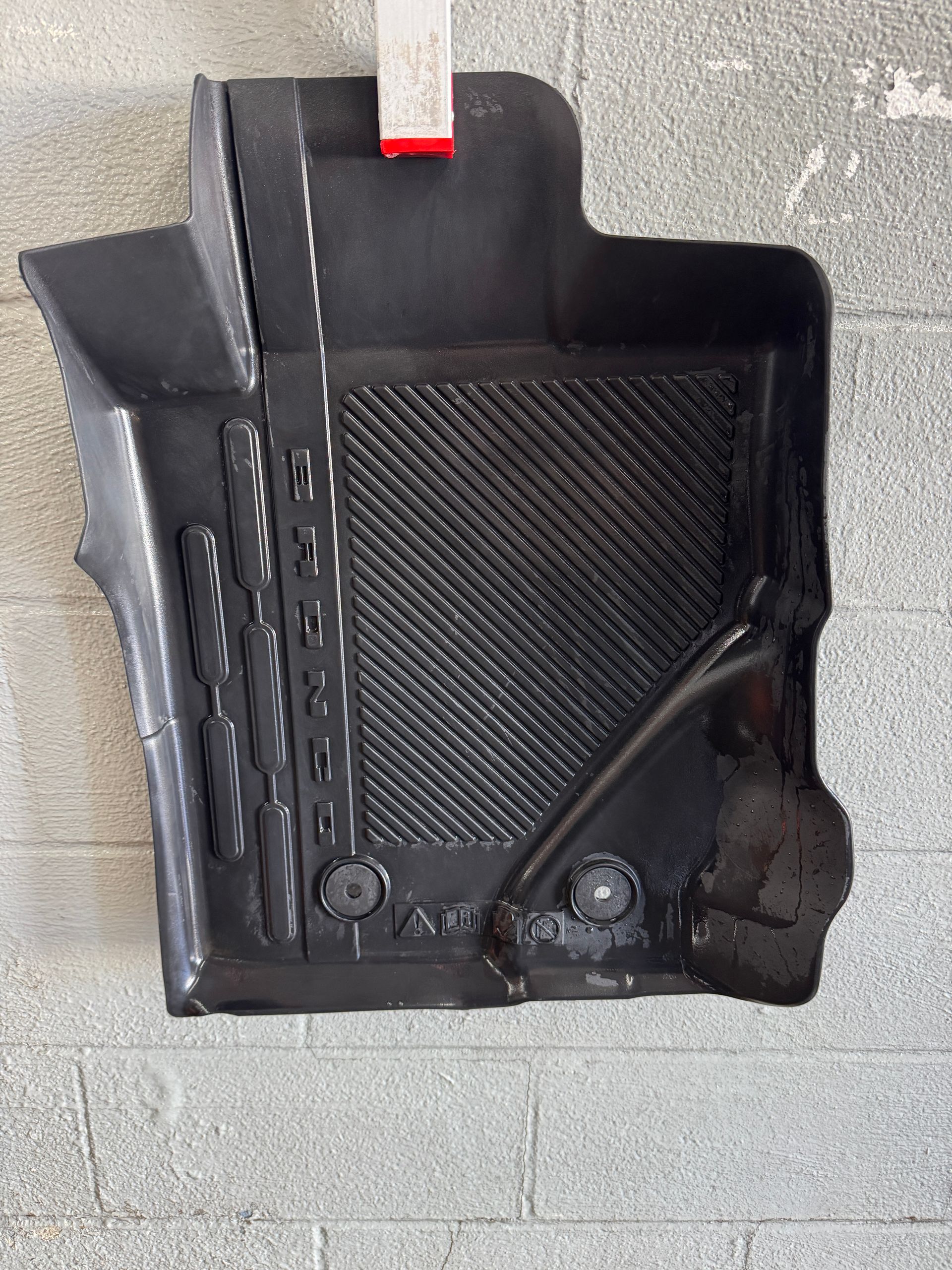 Black rubber car floor mat against a textured gray wall, showing grooves and raised edges.