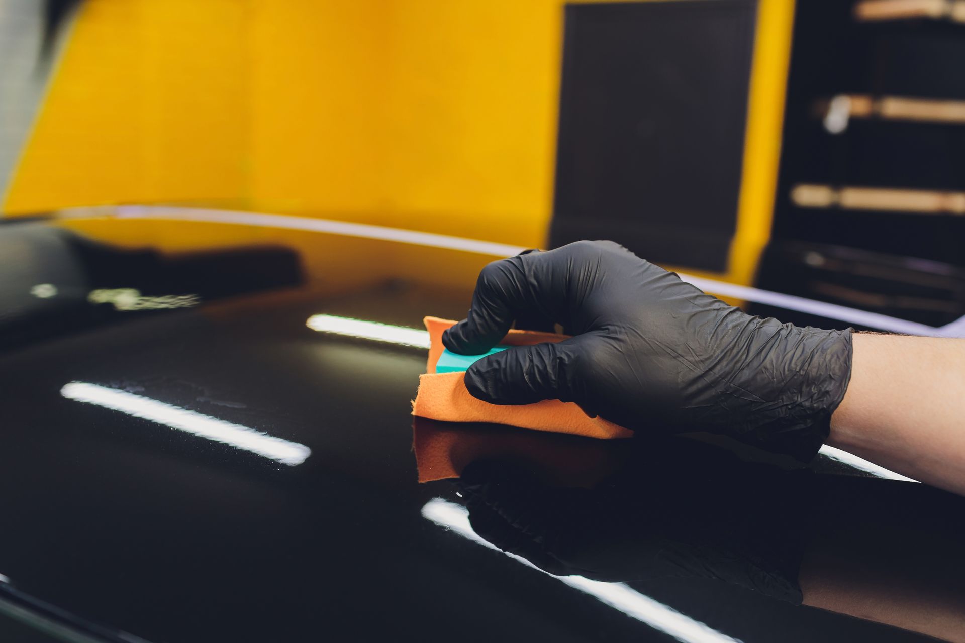 Gloved hand applying a ceramic coating to a shiny black car hood with a sponge.