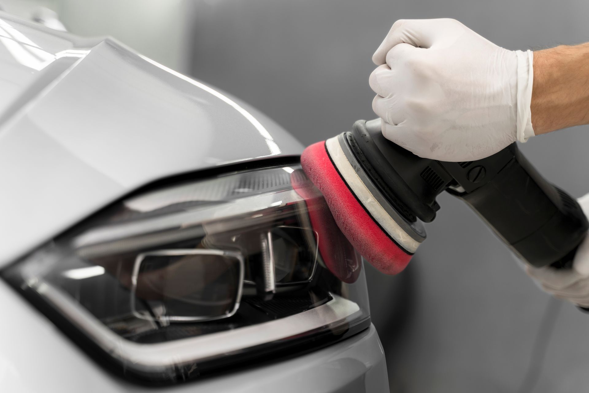 A gloved hand uses a polisher on a car headlight, restoring its clarity.