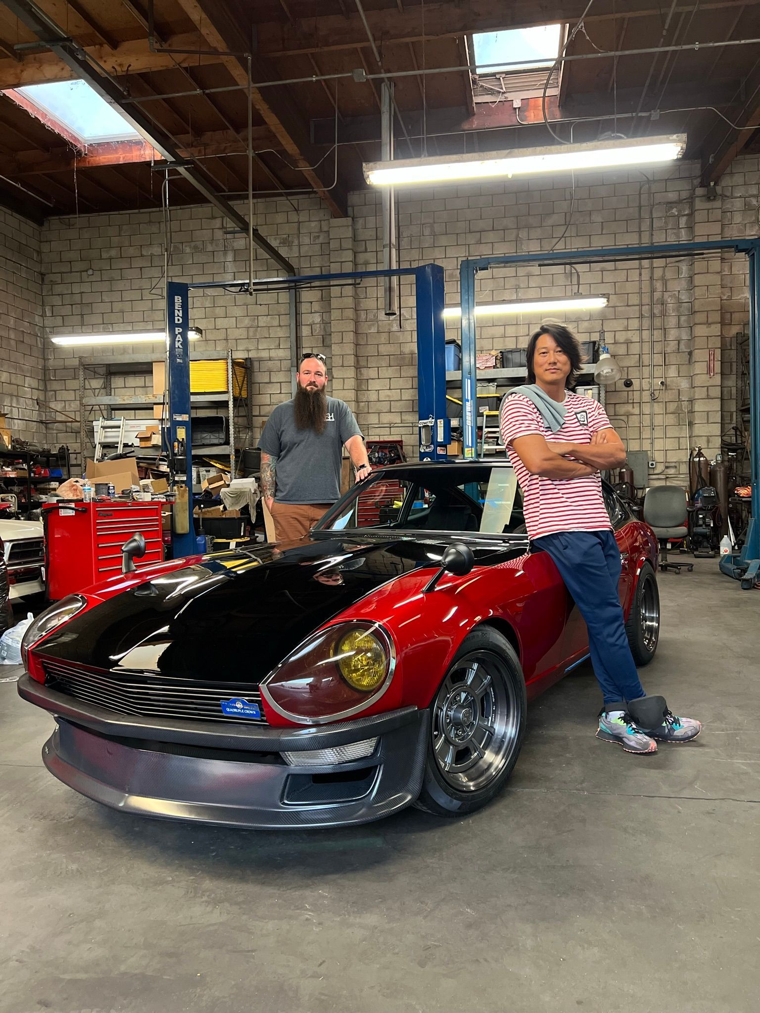 Two men pose with a custom red and black sports car in a garage.