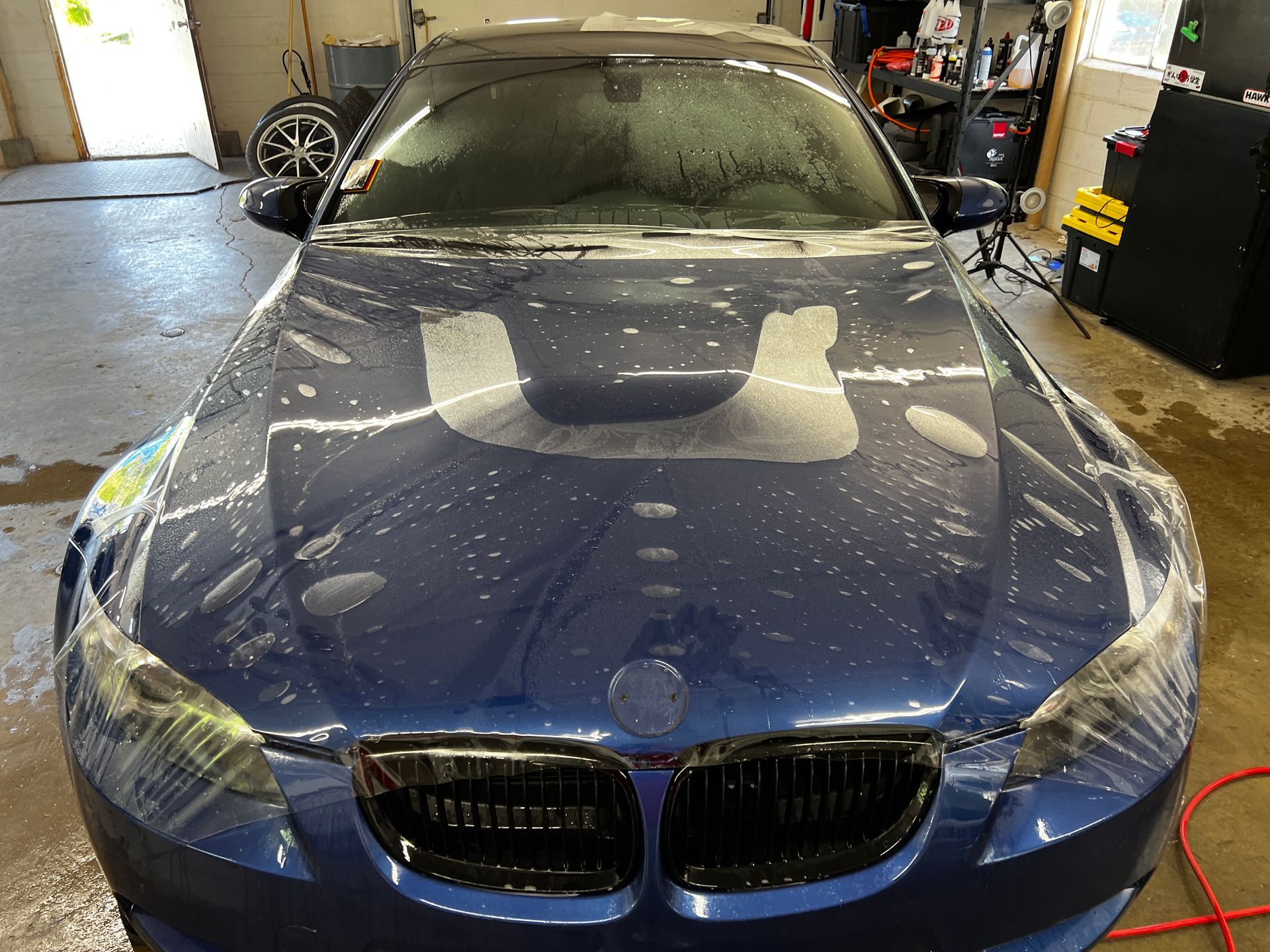 Blue car with wet hood; windshield and hood are being prepared for a film application in a garage setting.