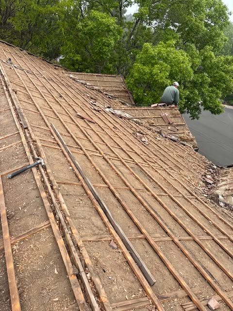 Damaged roof with exposed wooden slats and a person working near the edge by trees