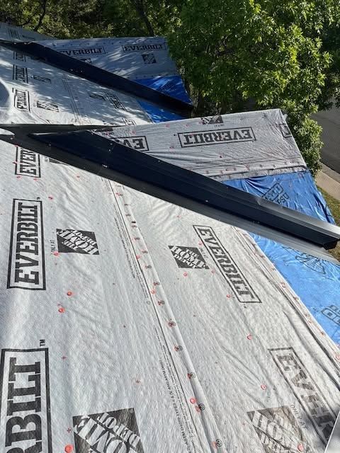 Roof under construction with gray Everlast underlayment, blue tarp, and trees nearby