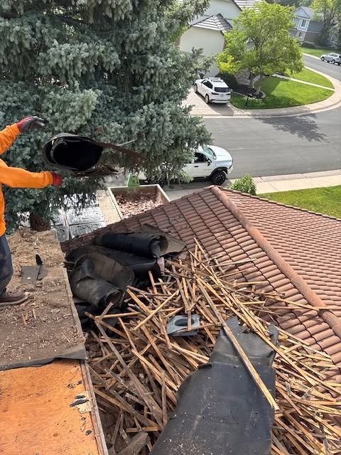 Worker on roof clearing black debris and twigs near a chimney on a red-tile roof, with a suburban street below.