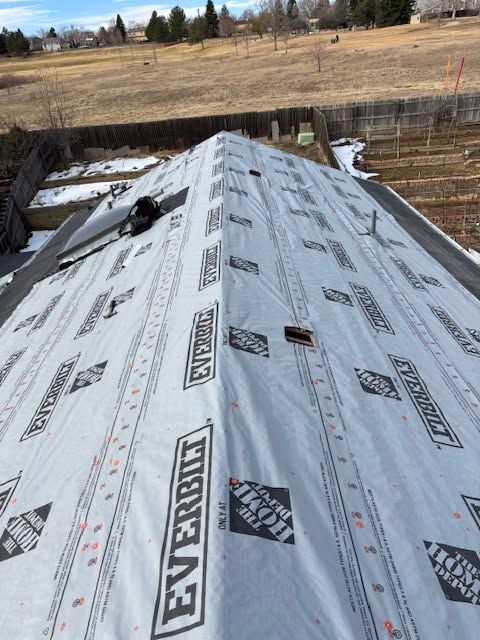 Roof under construction covered with gray underlayment, viewed over a fenced yard and open field.
