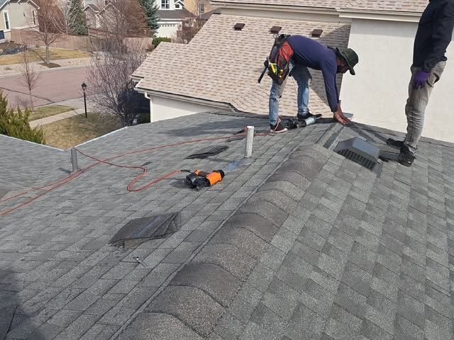 Roofers installing shingles on a gray roof, with tools near a vent stack.
