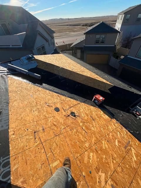 Roofer’s view of a steep shingle roof under construction with a tape measure and neighboring houses below.