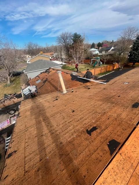 Rooftop with exposed plywood and scattered debris, damaged section near center, neighborhood trees and houses beyond