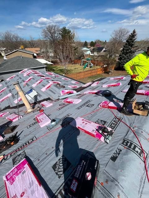 Roofer installing gray underlayment on a sloped roof, with tools and packaging scattered around.
