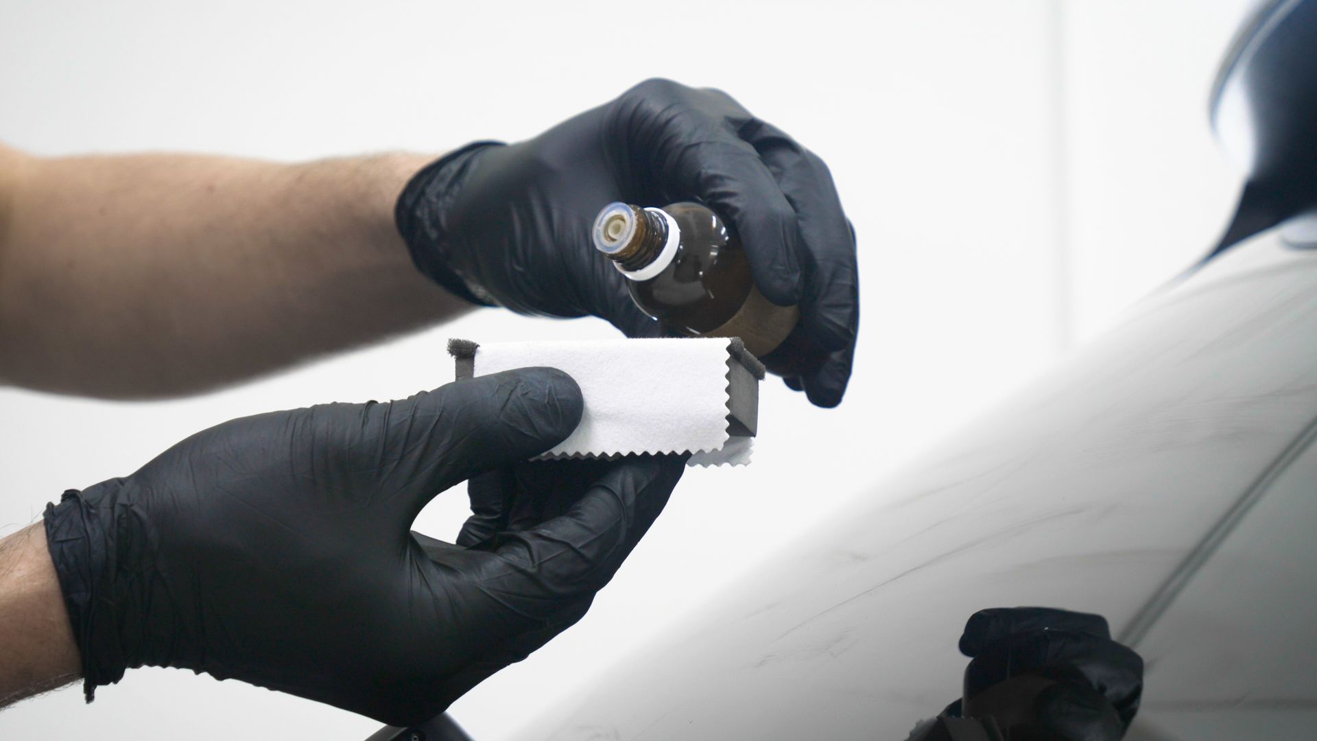 Person in black gloves pouring liquid from a bottle onto an applicator; near a white car panel.