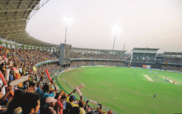 Crowded cricket stadium with fans cheering as players stand on the green field under bright lights