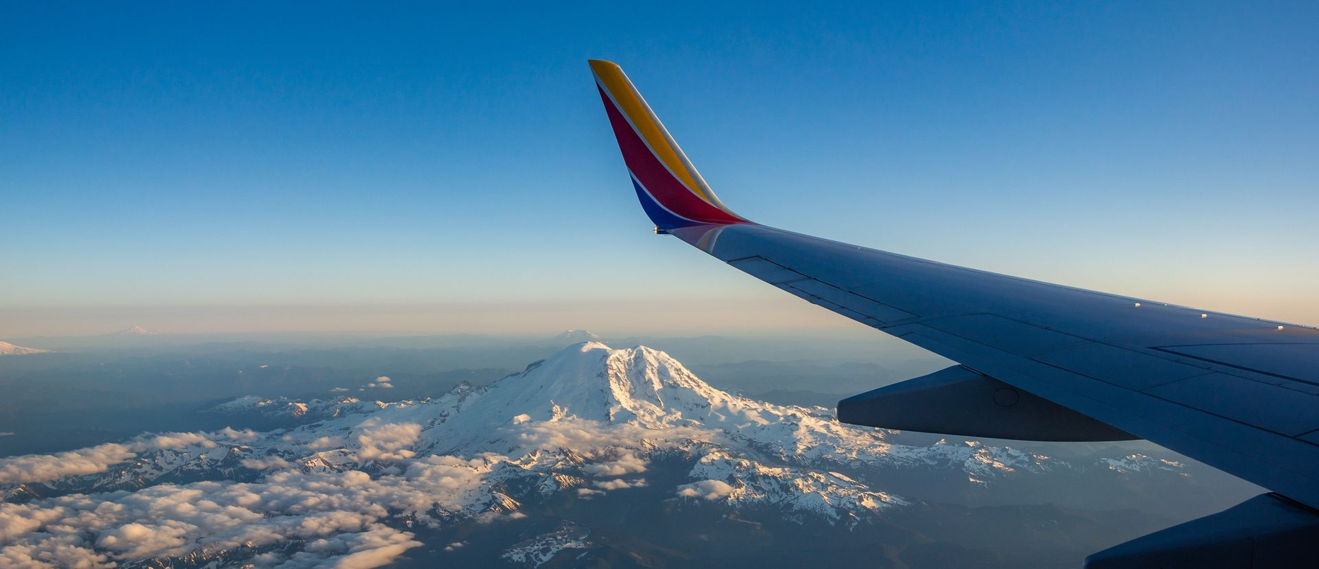 View from airplane window over snowy mountains at sunset, with wing and blue sky