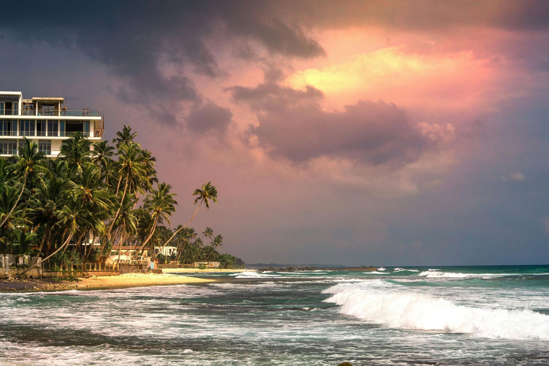 Tropical beach at sunset with palm trees, waves, and a coastal resort under dramatic pink clouds