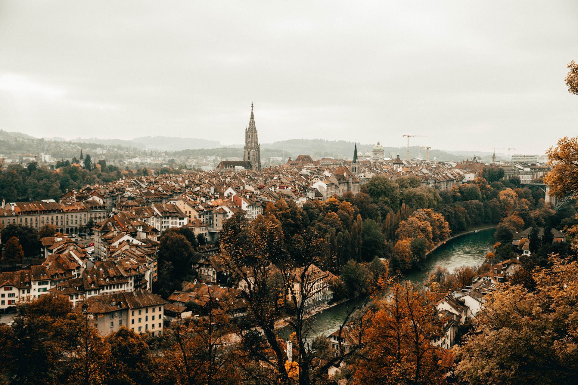 Panoramic cityscape of red-roofed buildings, a tall cathedral tower, and a river through autumn trees