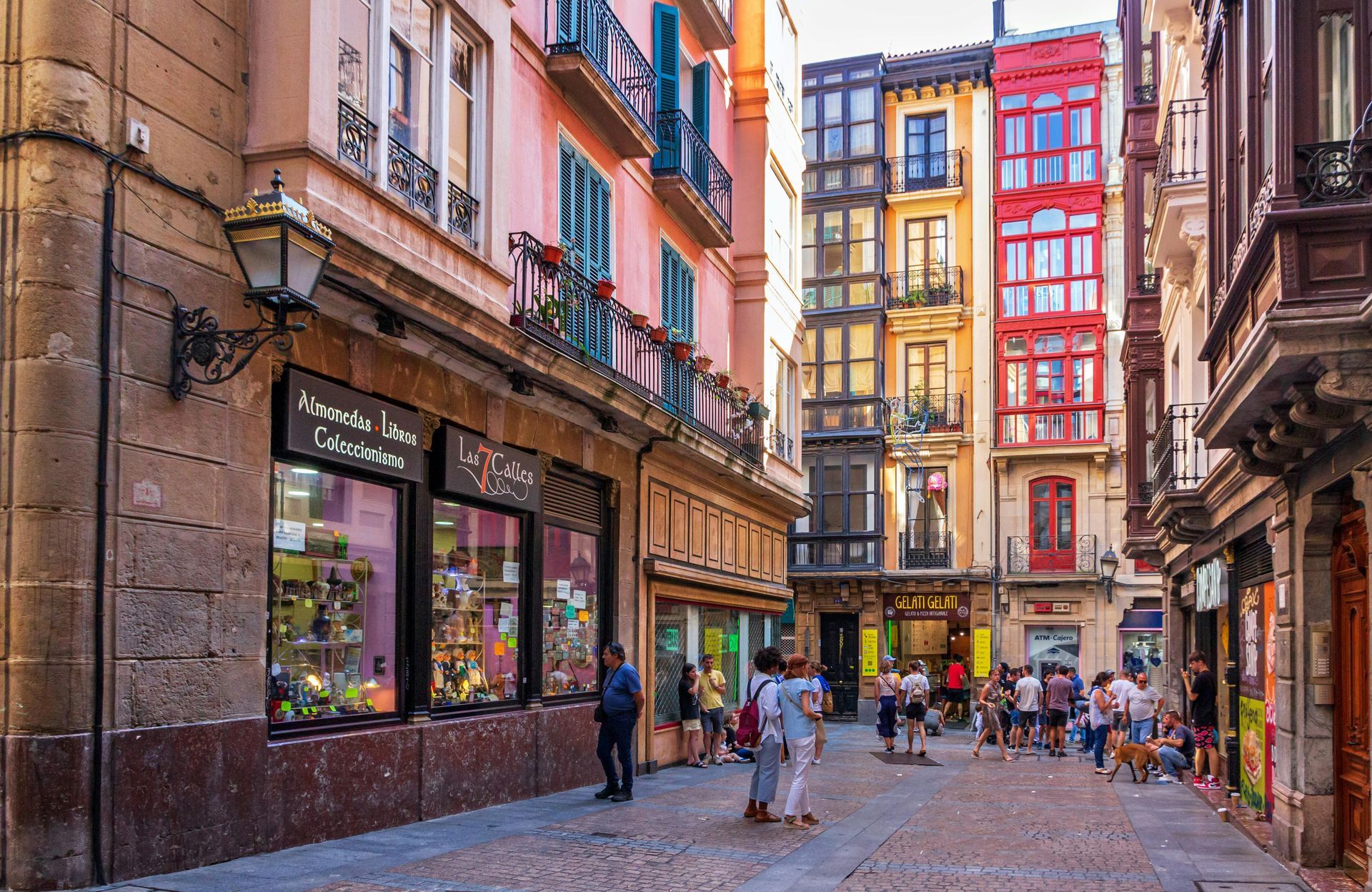 Narrow cobblestone street lined with colorful buildings and shops, with pedestrians walking under a bright sky