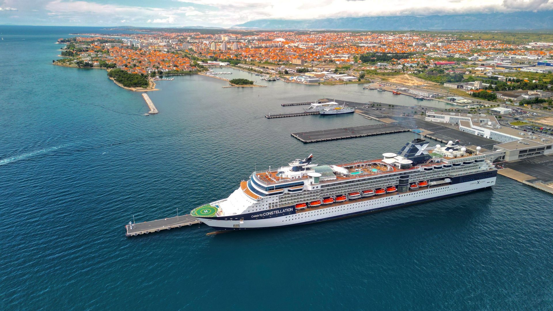 Cruise ship docked in a turquoise harbor beside a coastal city under a cloudy sky