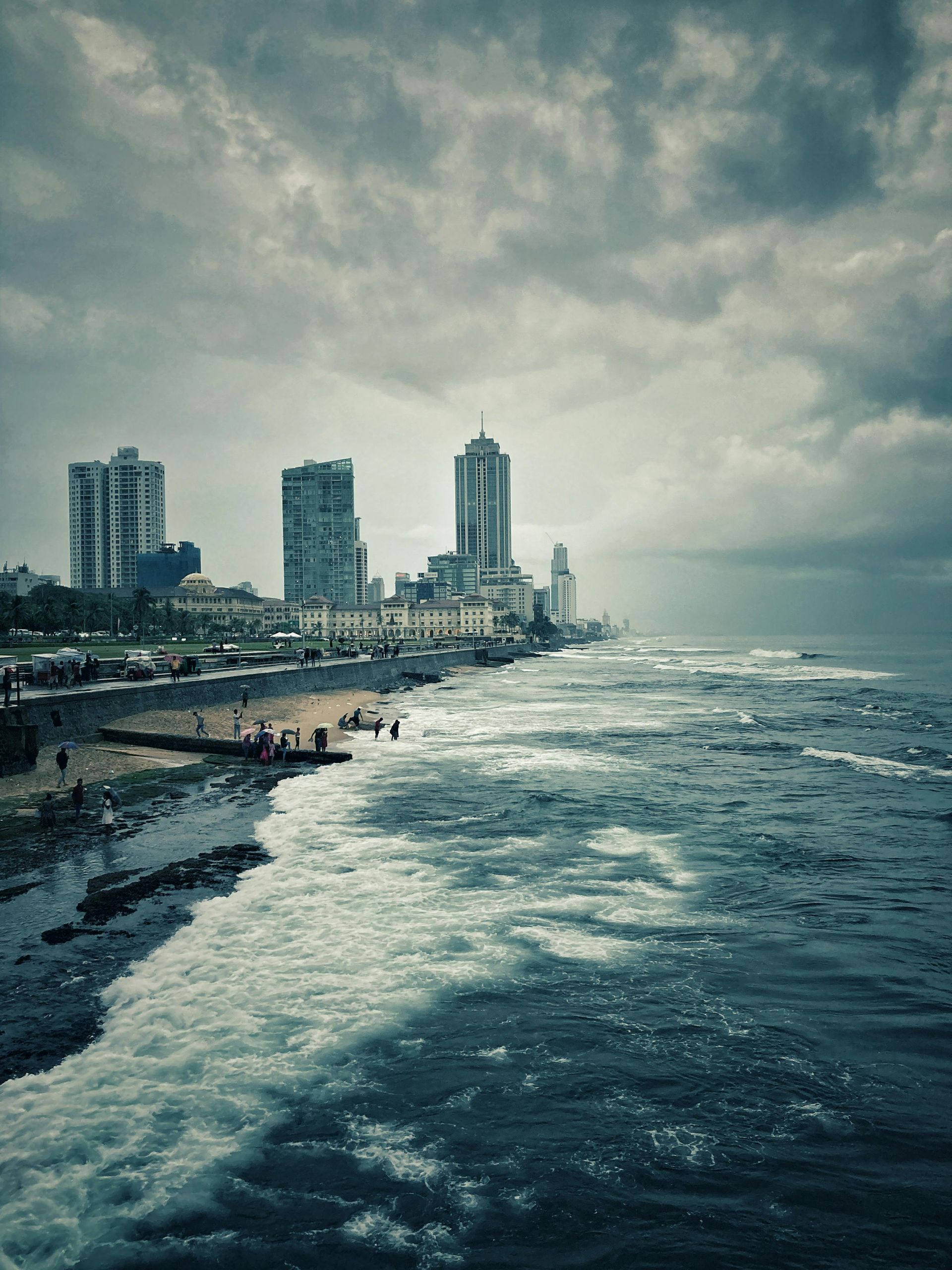 City skyline along a stormy coastline; waves crash on the shore, dark clouds overhead.