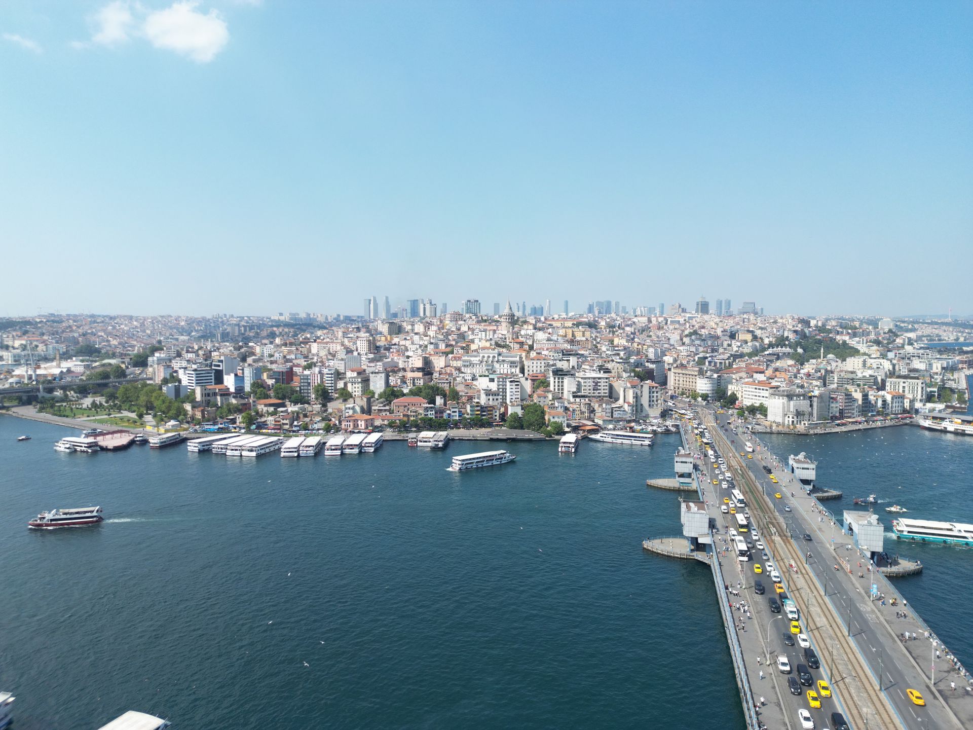 Aerial view of a coastal city with a bridge, harbor boats, and dense skyline under a clear blue sky