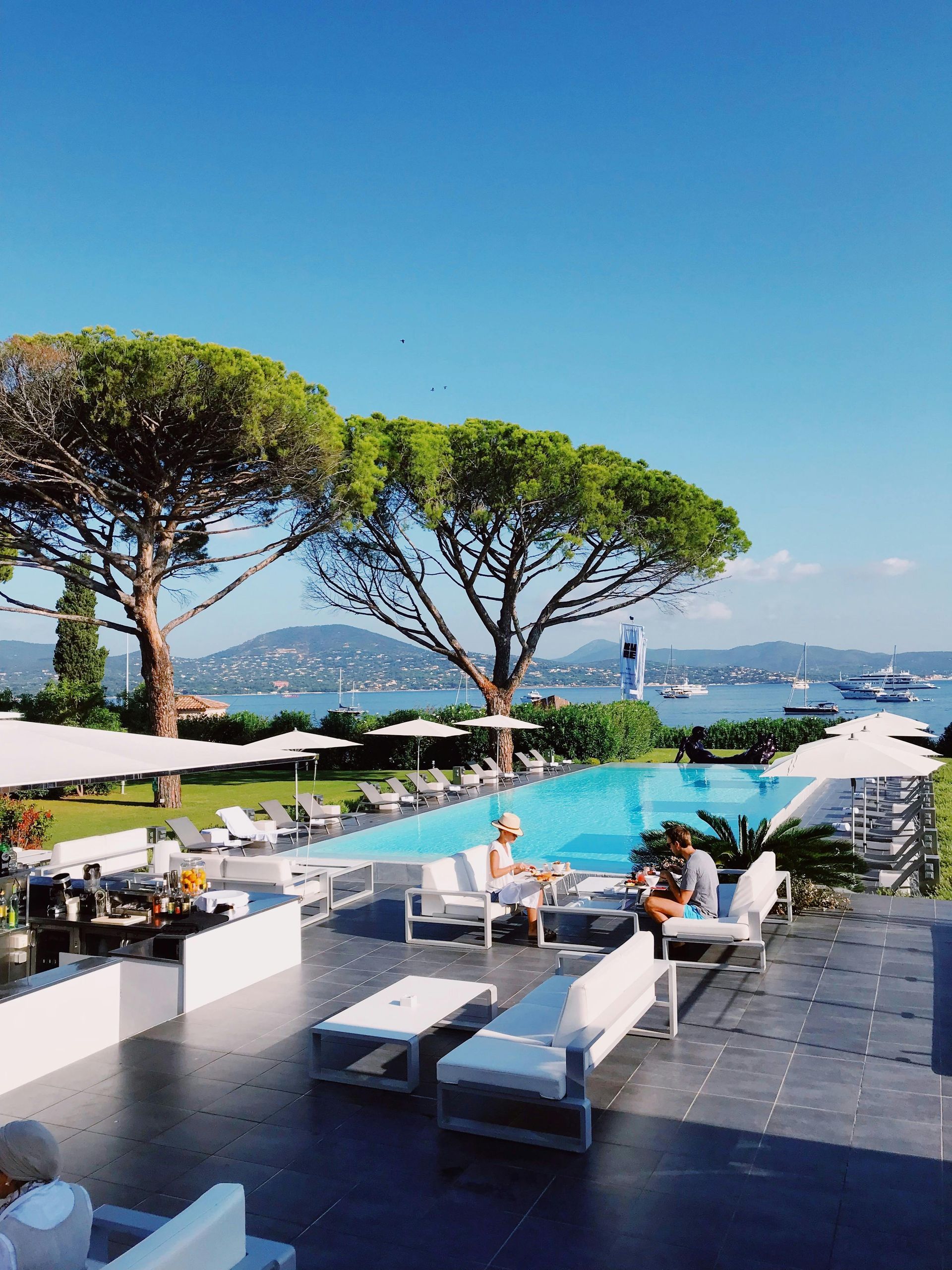 A pool overlooking a bay with people lounging on white furniture under blue skies.