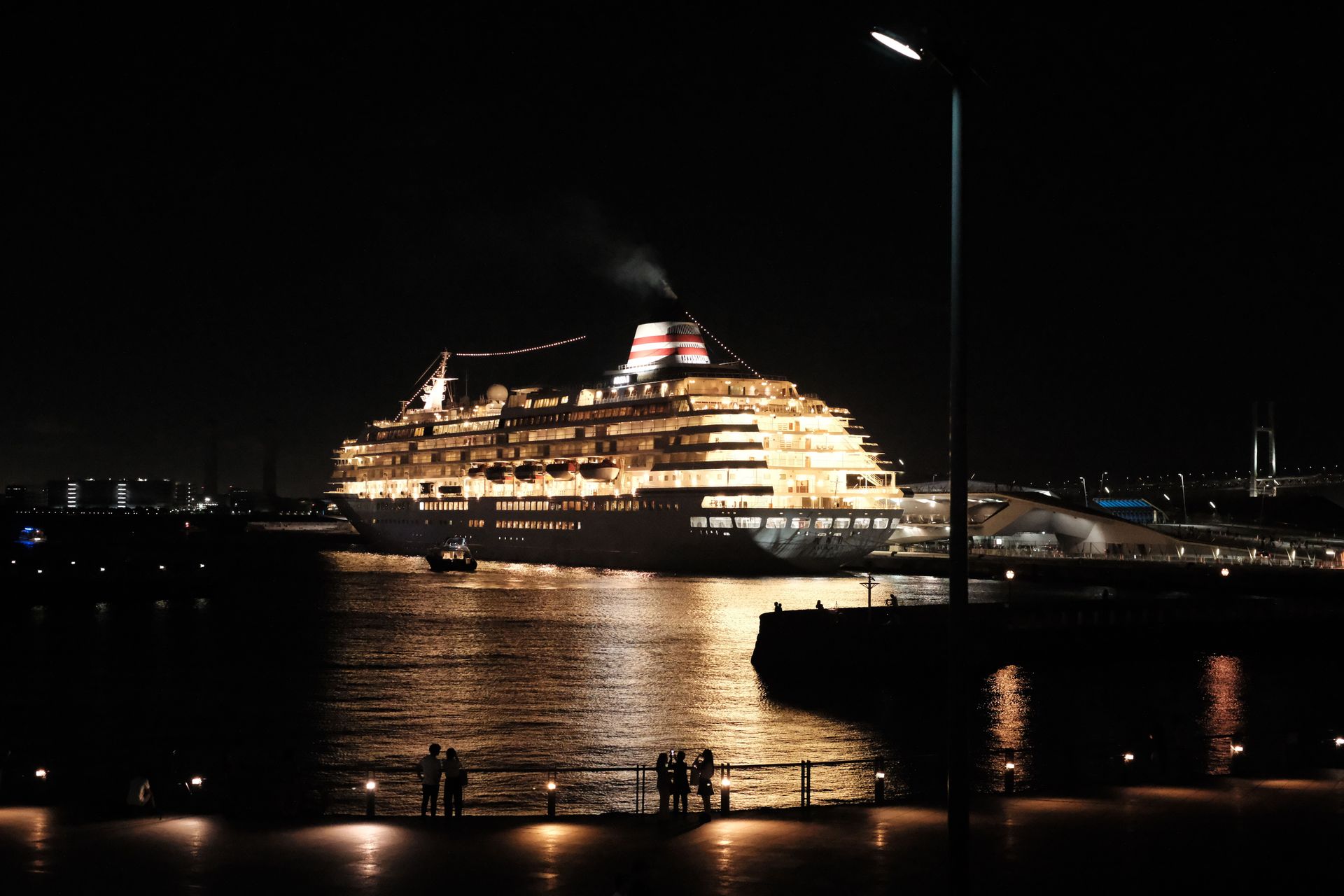 Lit cruise ship glowing at night on calm water beside a harbor streetlight