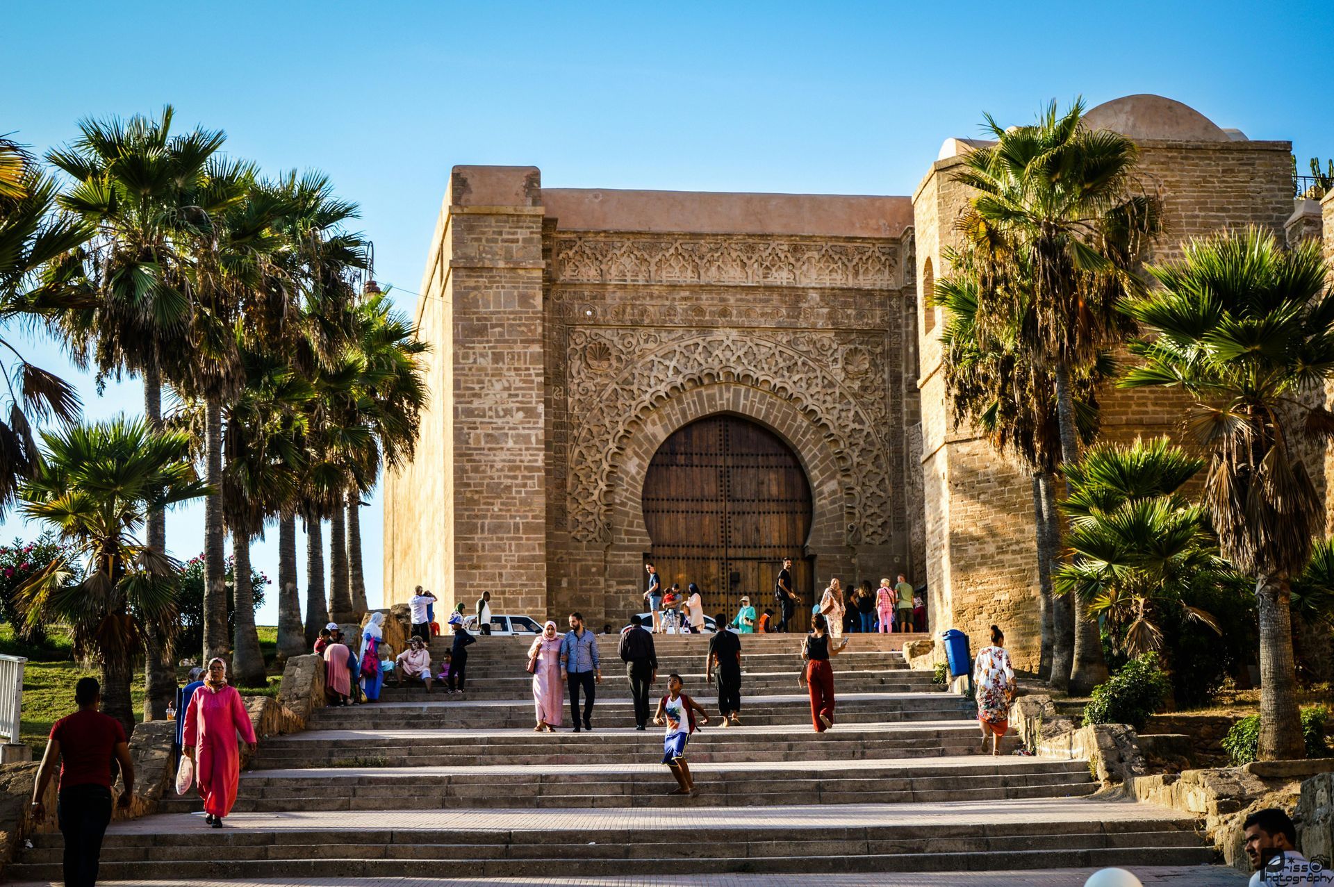 Stone mausoleum with arched doorway, palm trees, and people ascending wide steps under a blue sky
