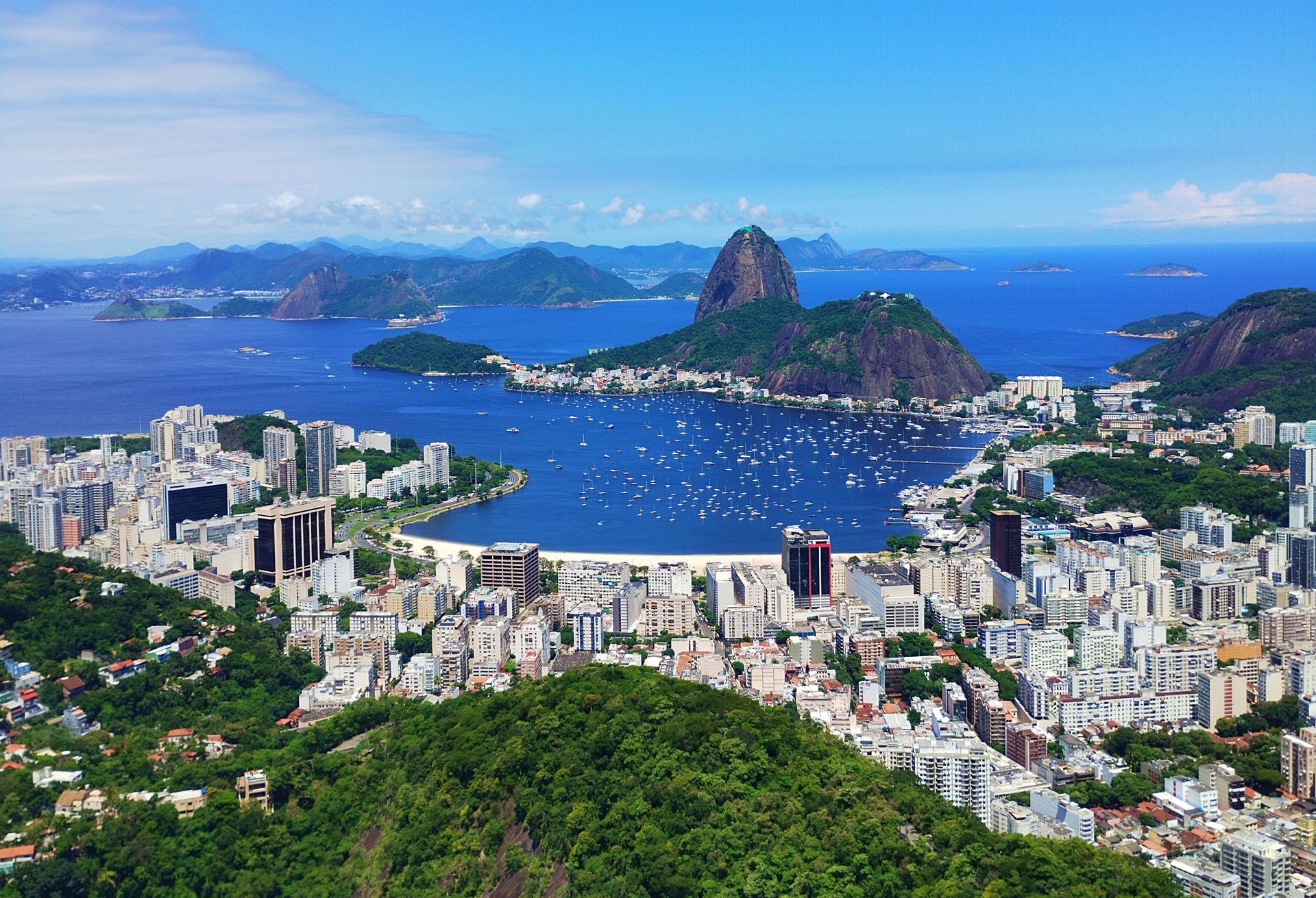 Panoramic cityscape with a bay, green hills, and rocky islands under a blue sky