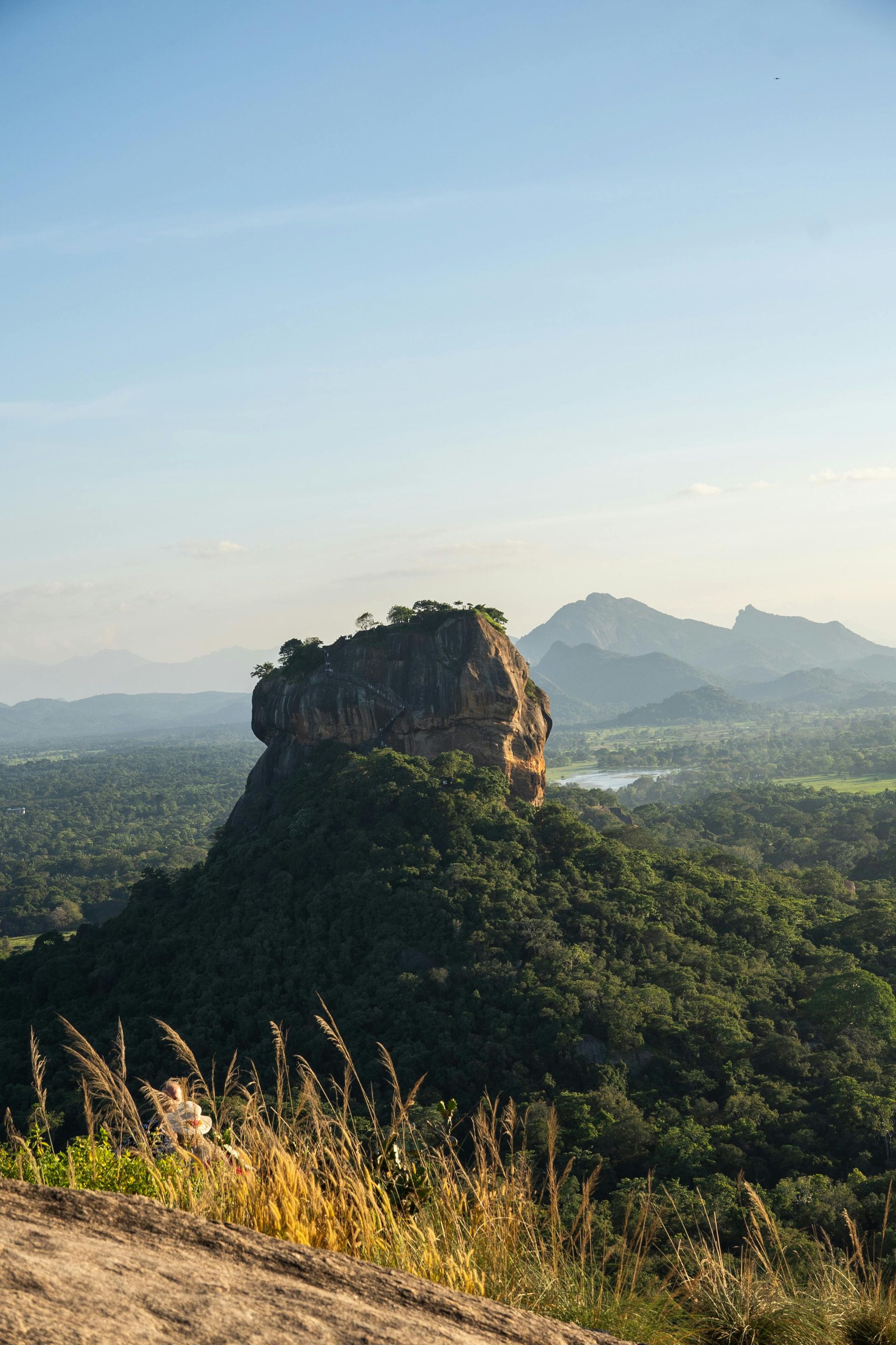 Sigiriya rock rising above a lush green valley at sunrise, with distant mountains and a clear blue sky
