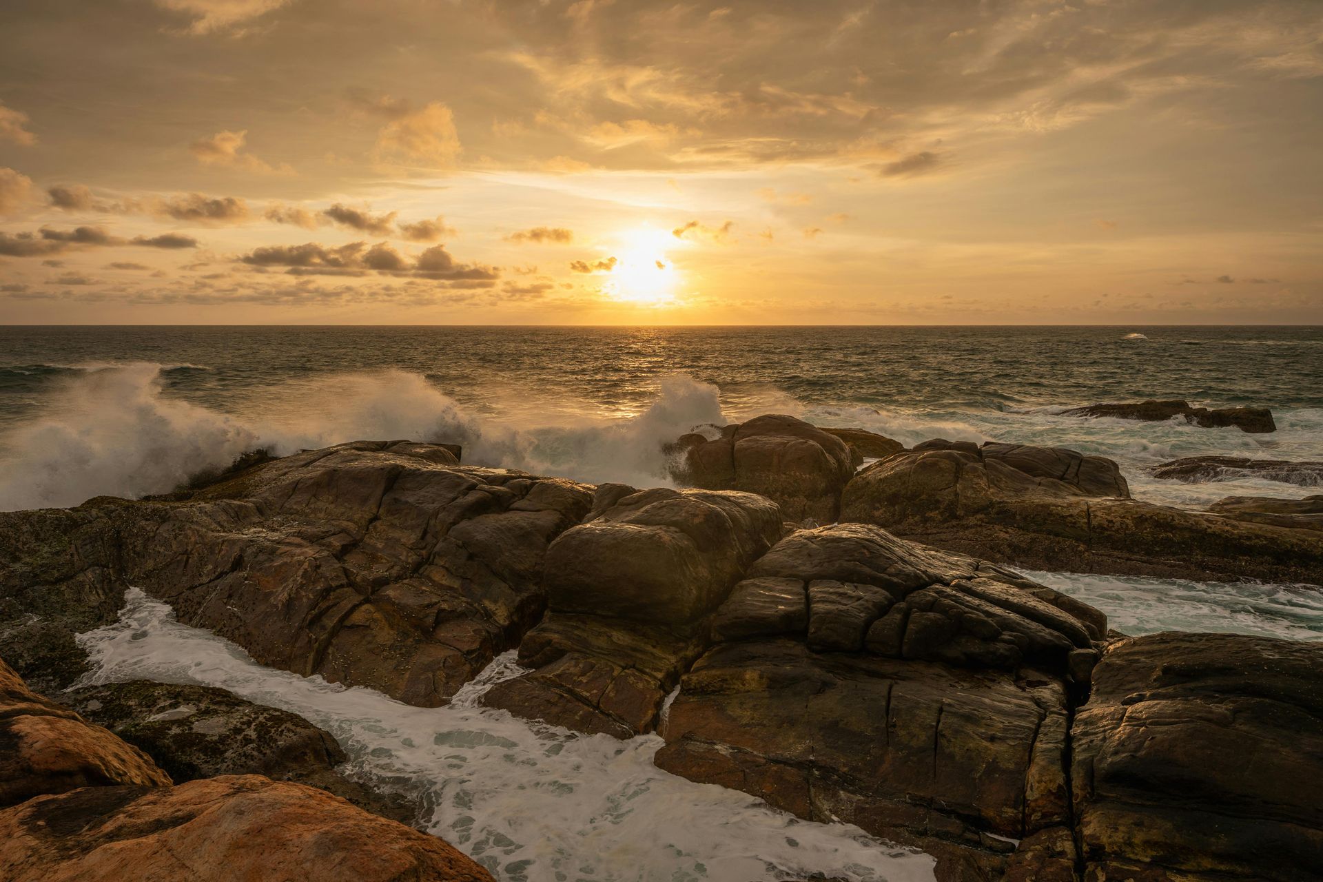 Sunset over rocky shoreline with waves crashing and golden light on the water