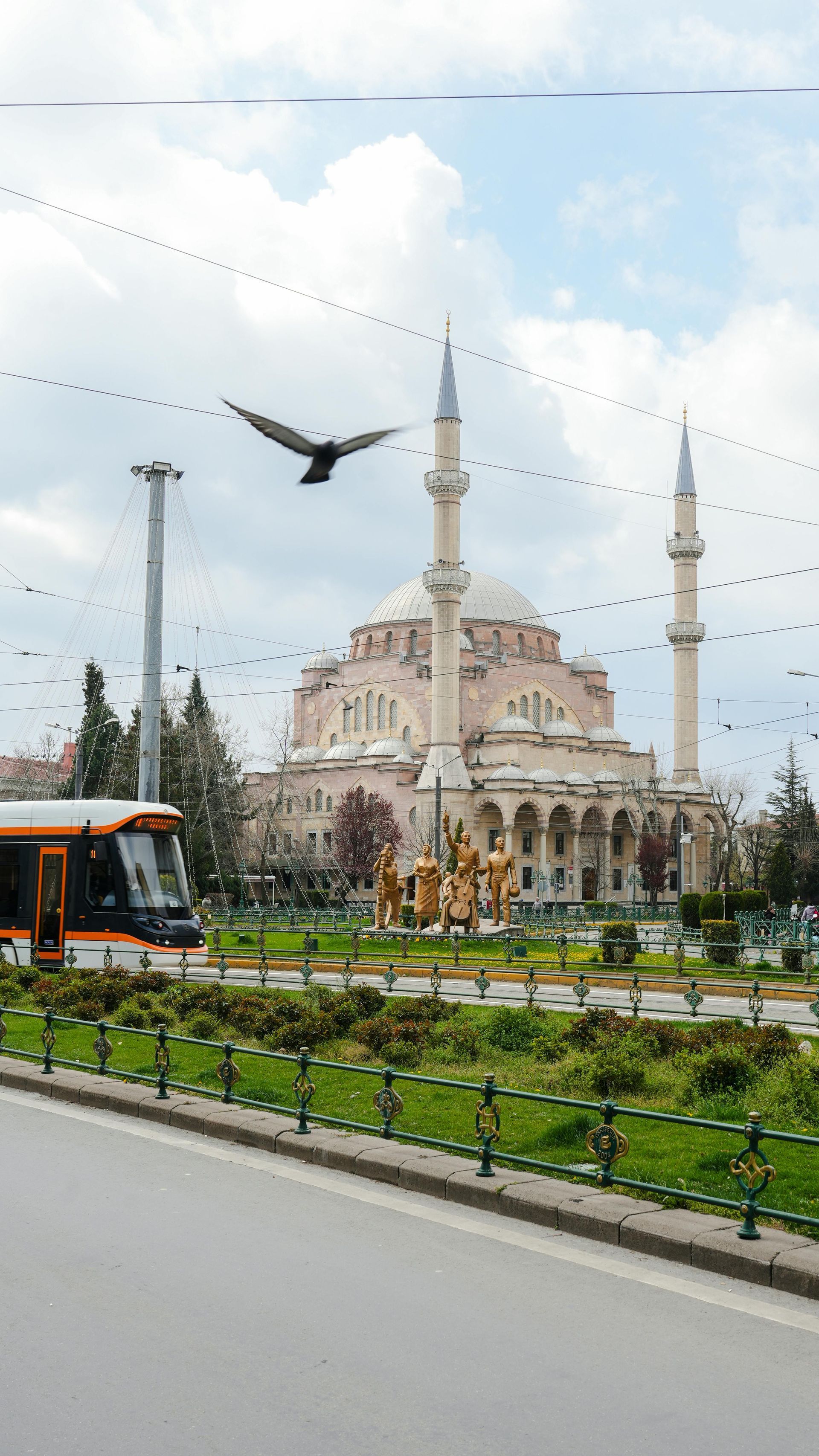 Mosque with minarets and a domed roof beside a road, with a tram and landscaped median in front