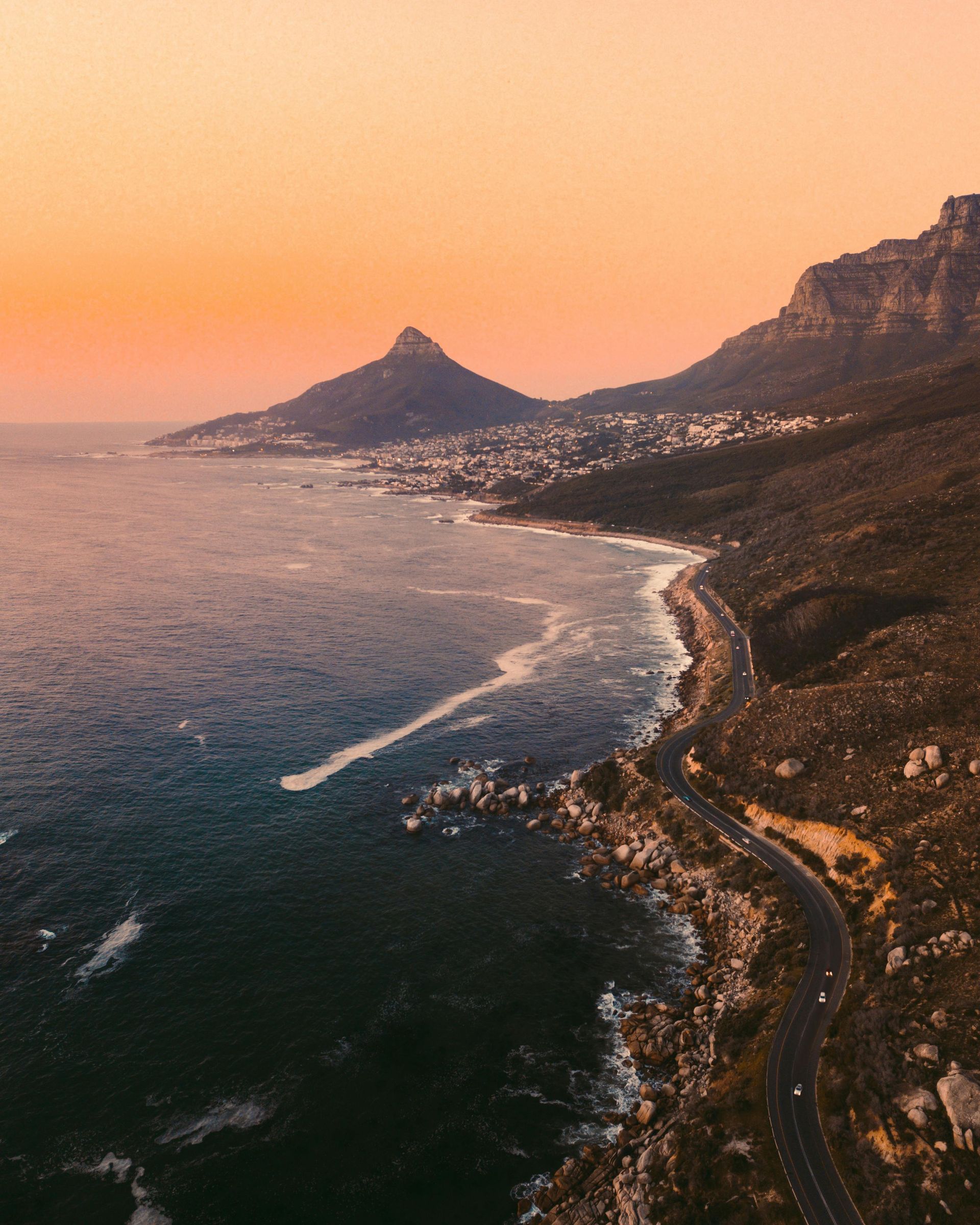 Coastline at sunset with a mountain, road, and buildings. Orange sky over blue ocean.