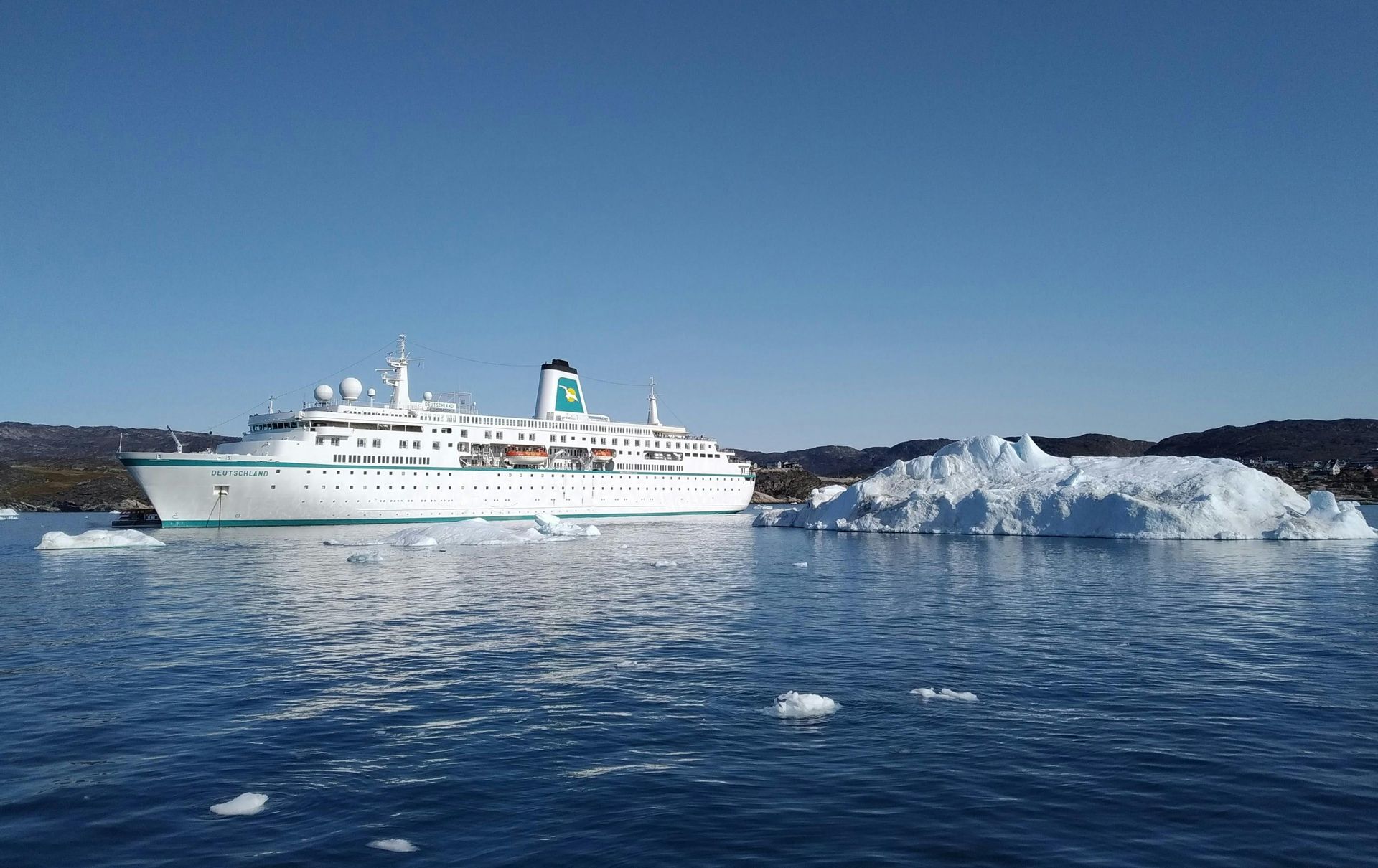 White cruise ship sailing beside an iceberg on calm blue water under a clear sky.