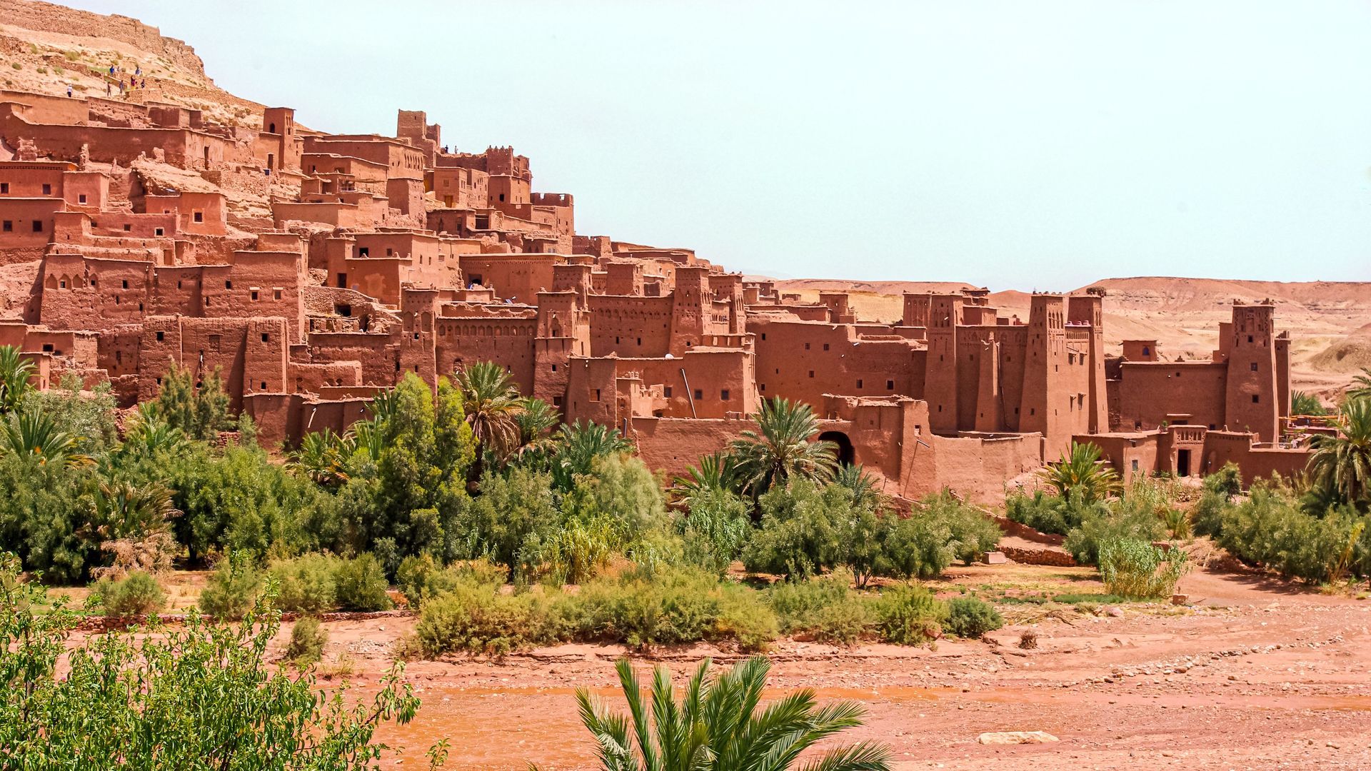 Red desert village with mud-brick houses beneath rocky cliffs and sparse greenery