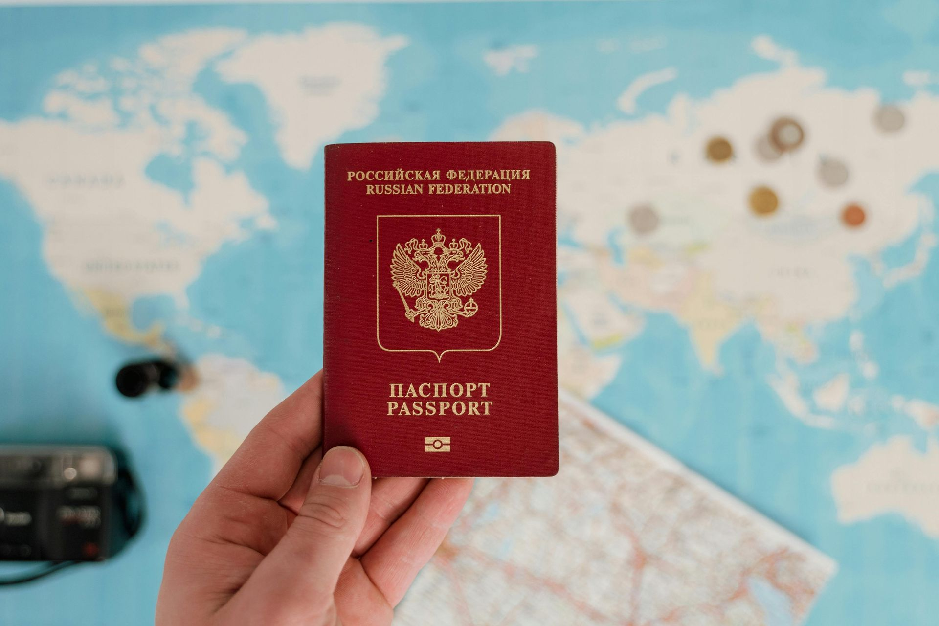 A hand holds a red Russian passport against a world map background with scattered coins and a camera nearby.