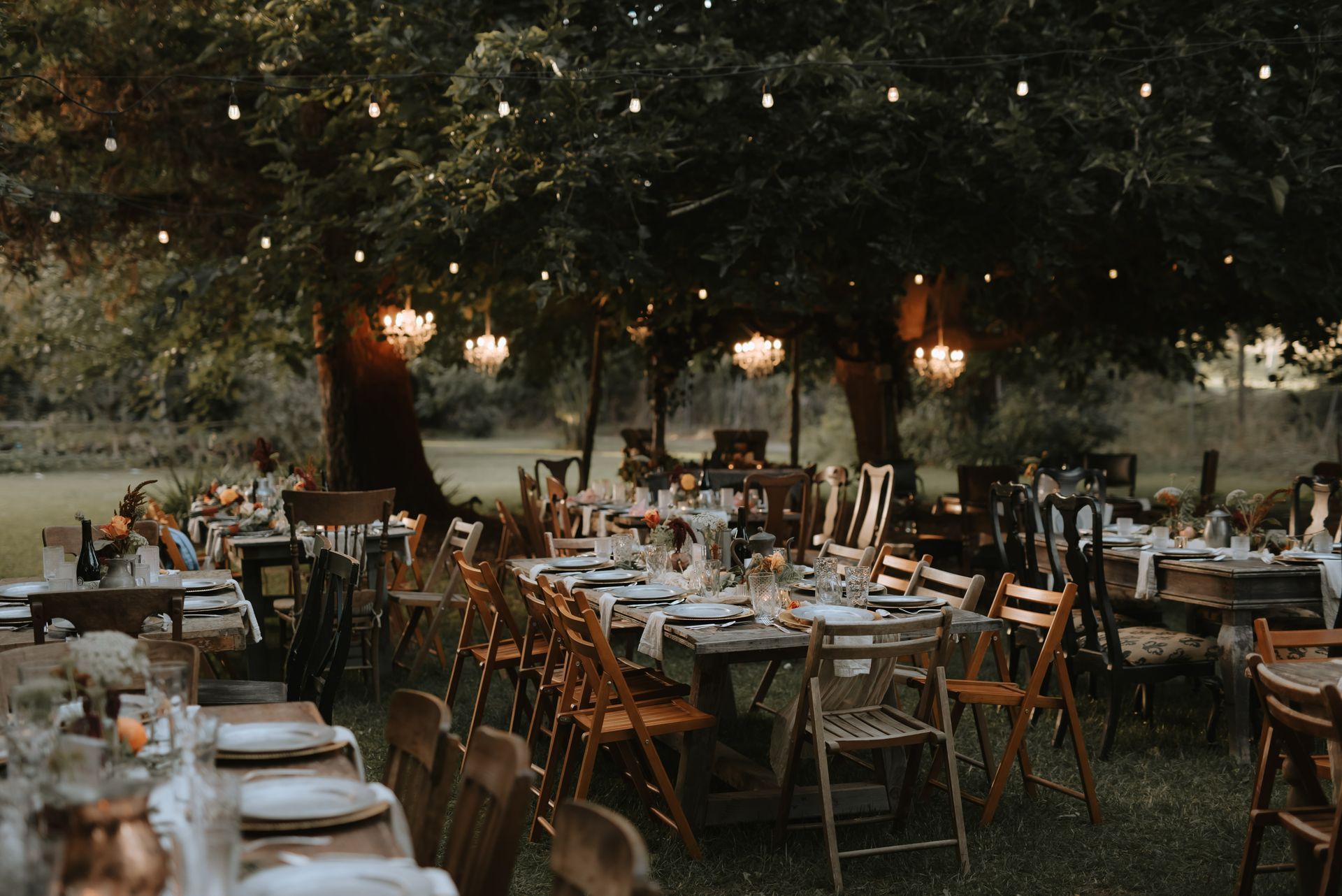 Outdoor reception tables set for a celebration, string lights and chandeliers illuminate the scene.