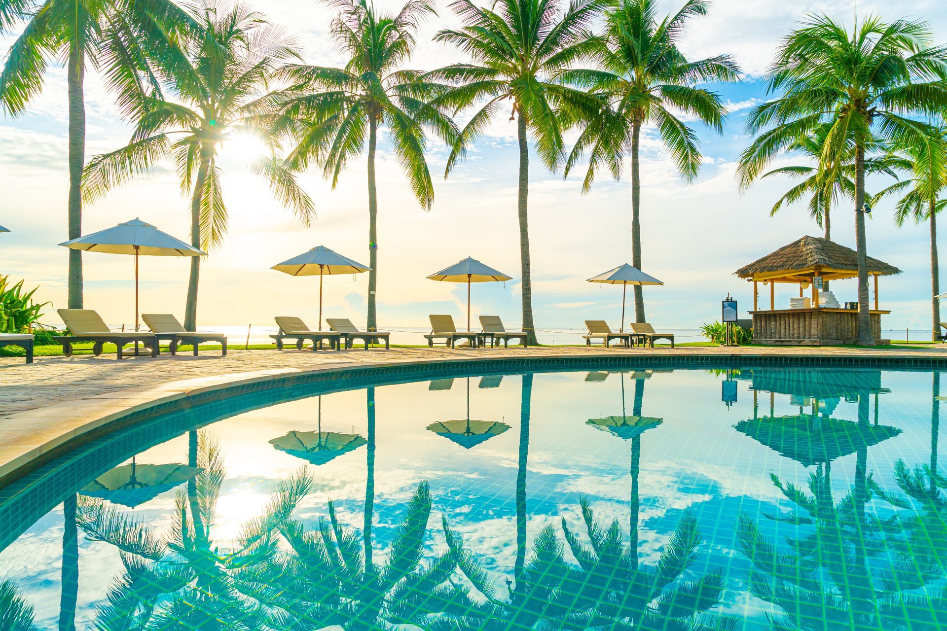 Poolside scene: Palm trees, lounge chairs, umbrellas, and a swim-up bar. The pool reflects the sky and trees.