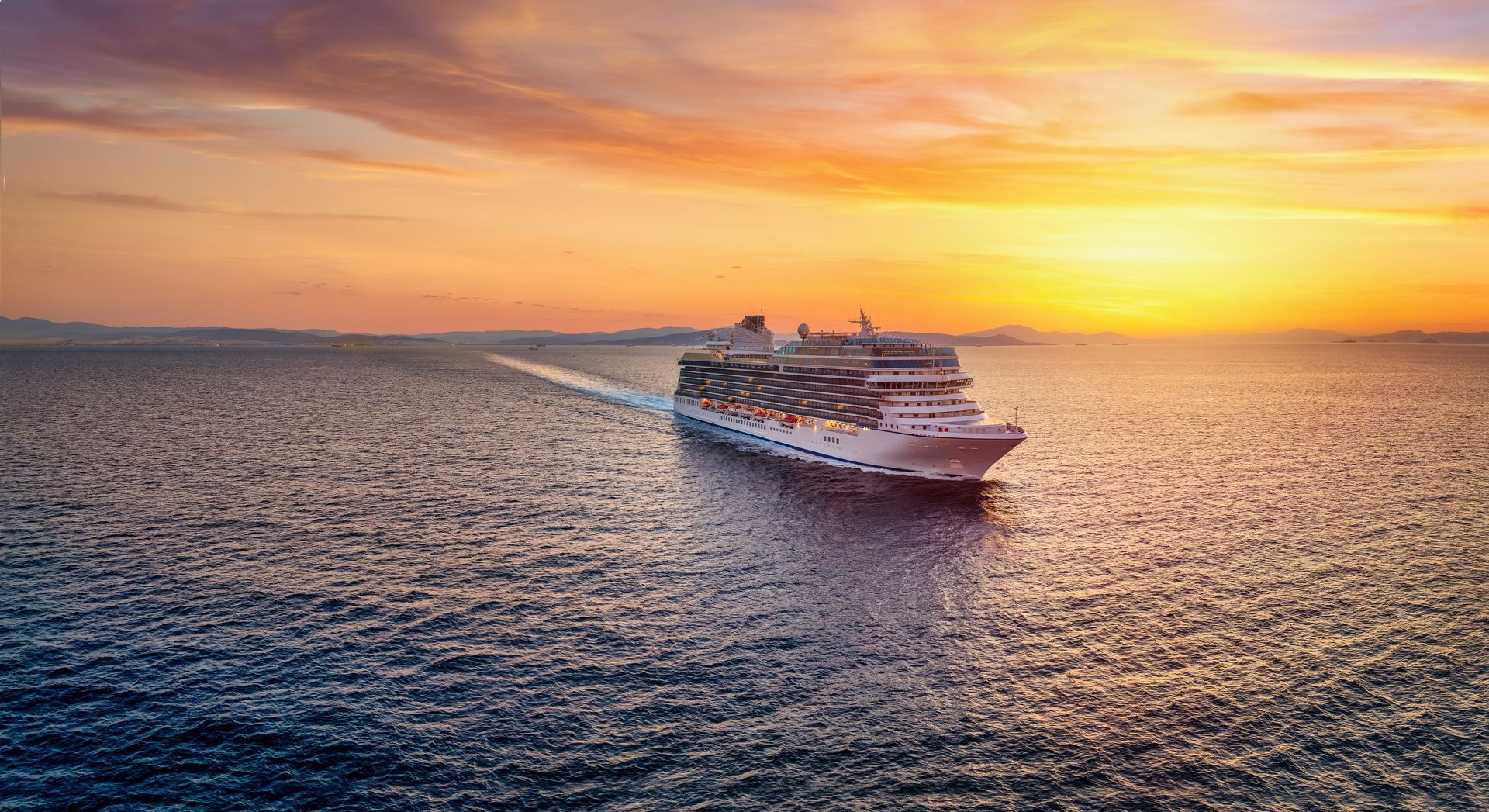 Cruise ship sailing on the ocean at sunset, with a warm orange sky and calm water.