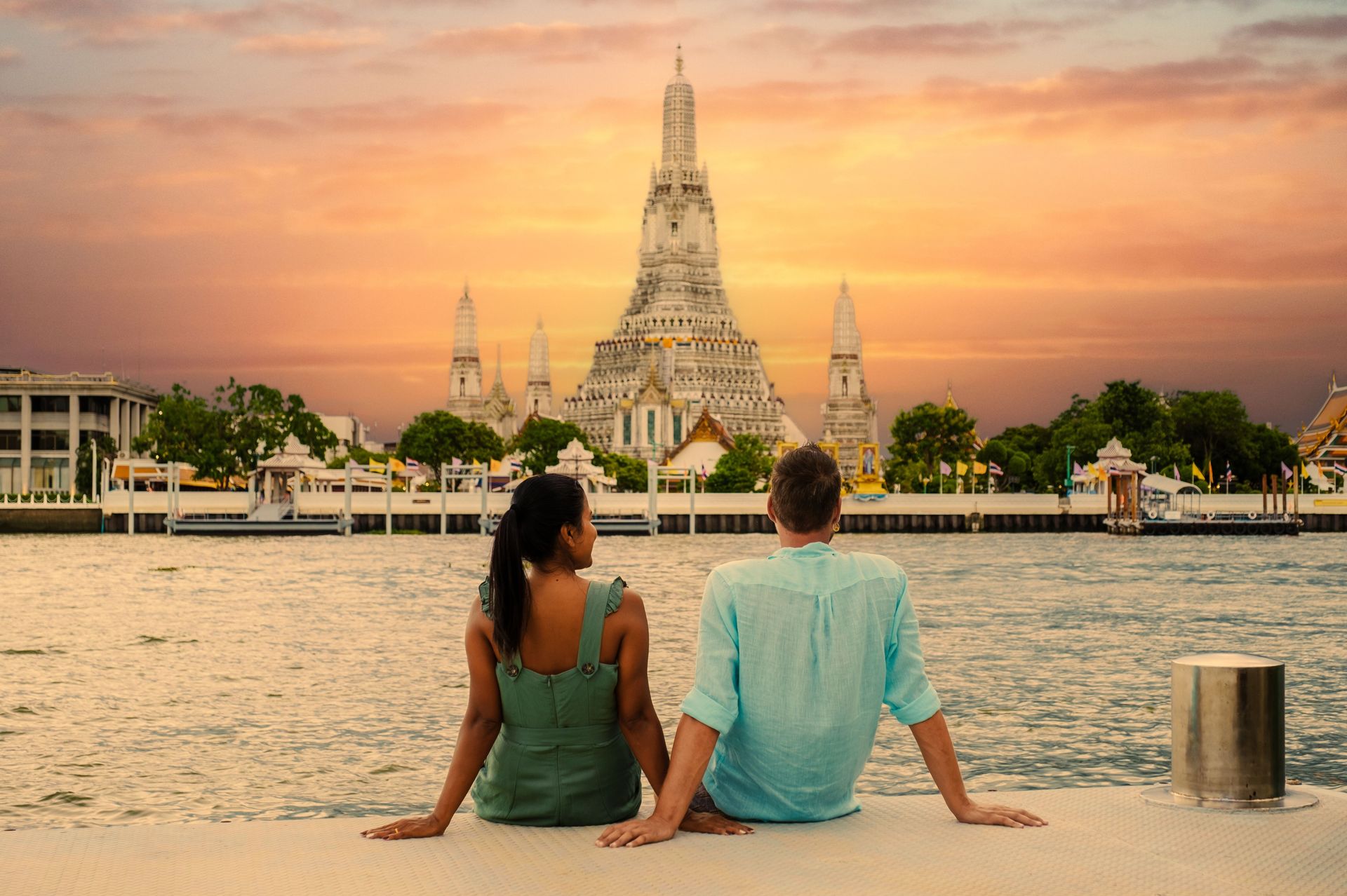 Couple sits overlooking a river at sunset, with a white ornate temple in the background.
