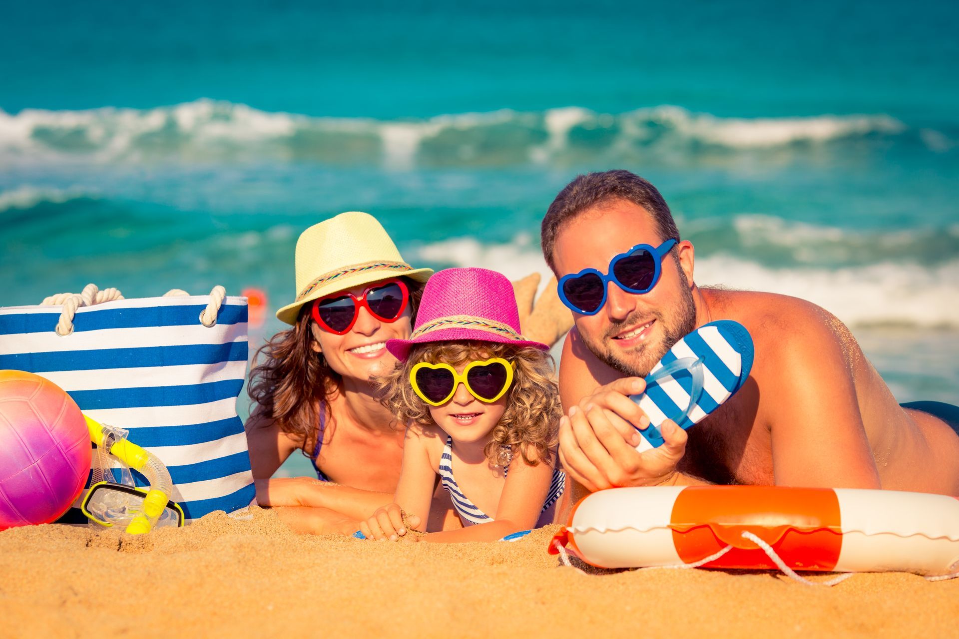 Family on beach: Woman, child, and man in sunglasses, hats, smiling, on sand near water, with beach bag and ball.