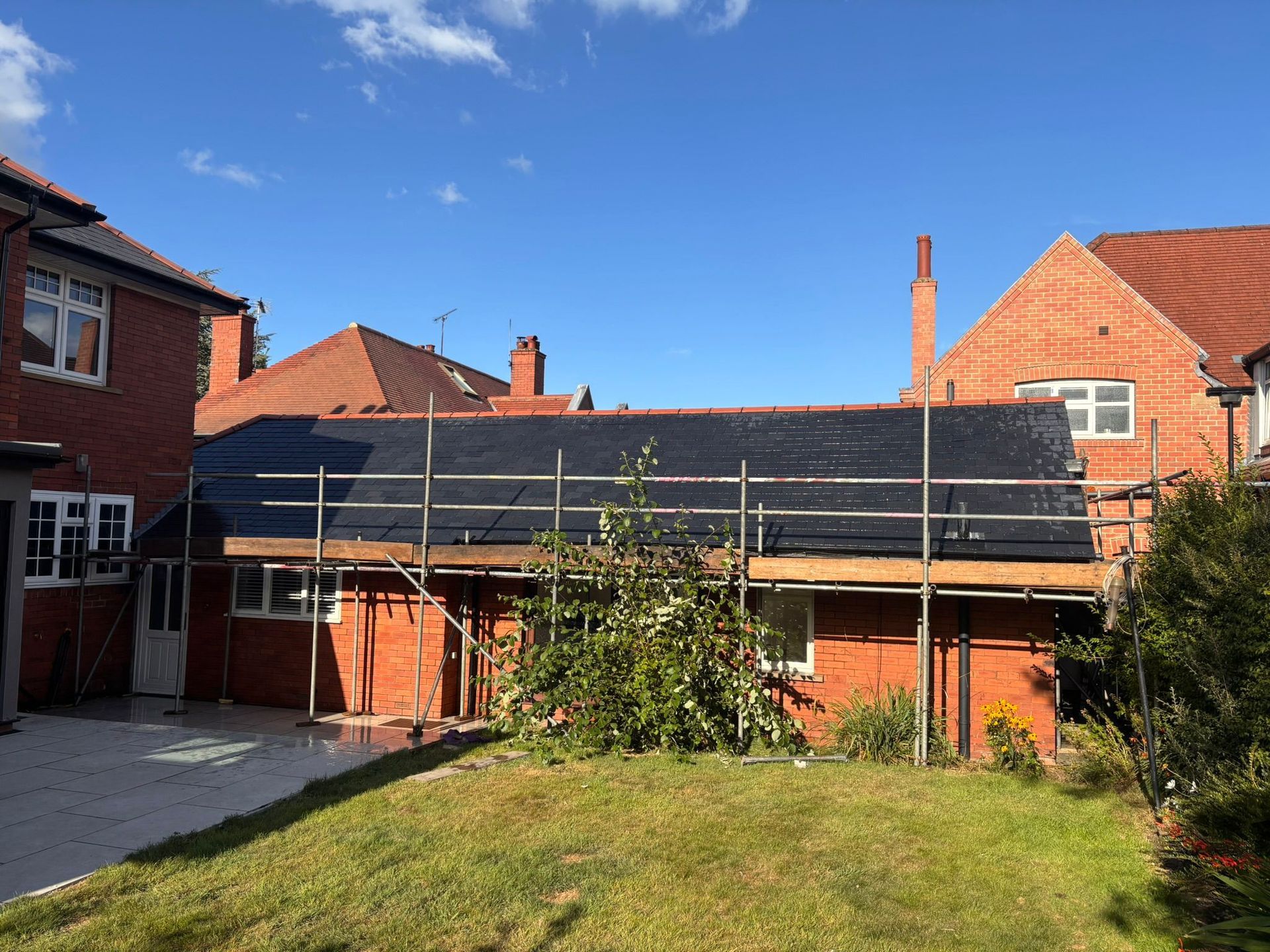 A rear view of a red-brick residential house under renovation, featuring a black-felted roof extension and metal scaffolding.